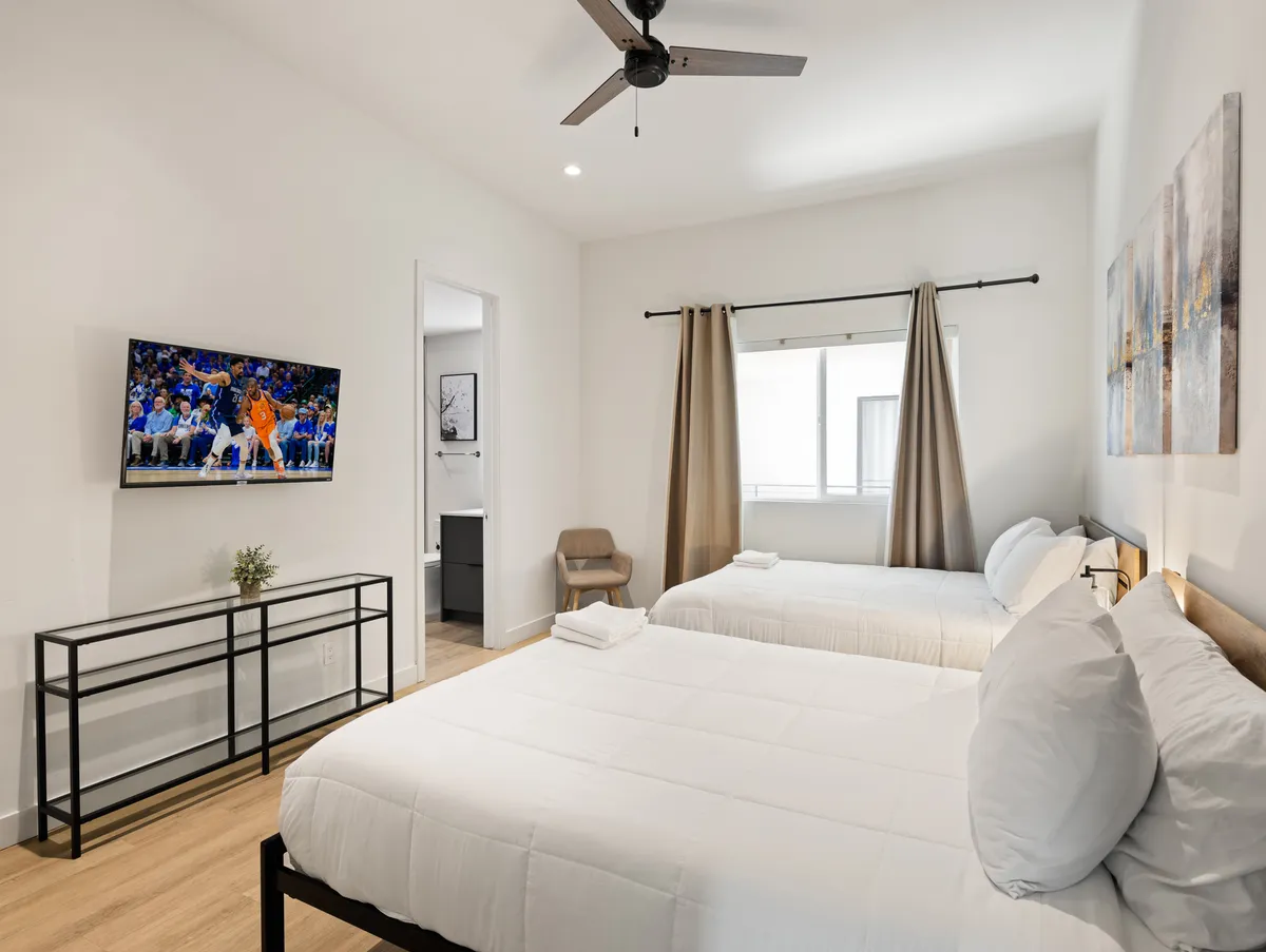 A cozy bedroom featuring two beds, a TV, and a window with light curtains. The overall mood is restful and inviting, perfect for unwinding after a day of activities or enjoying a family movie night.