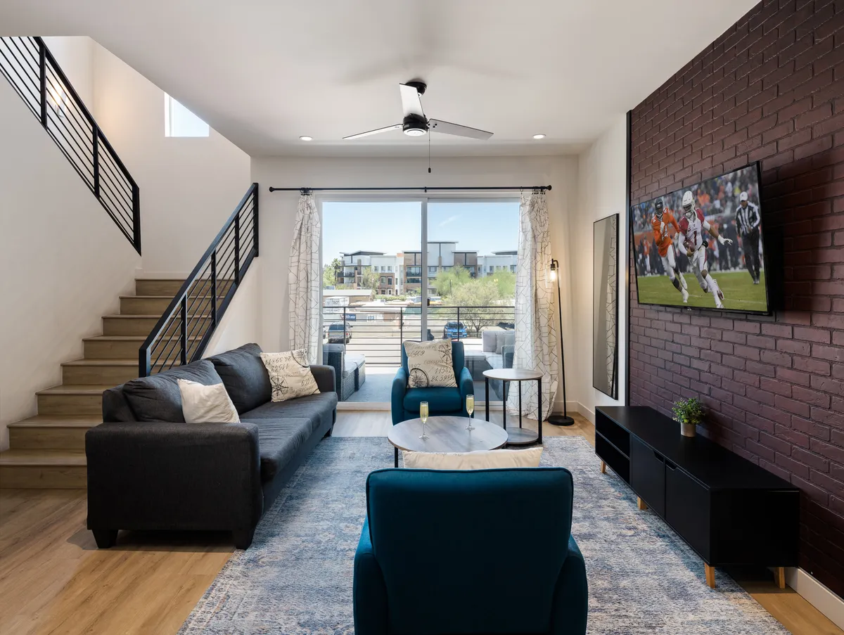 A bright, modern living room with a large window letting in natural light. A comfortable sofa faces a wall-mounted Smart TV, which is displaying a sports game. The space feels casual yet stylish, with ample room for relaxing or socializing.
