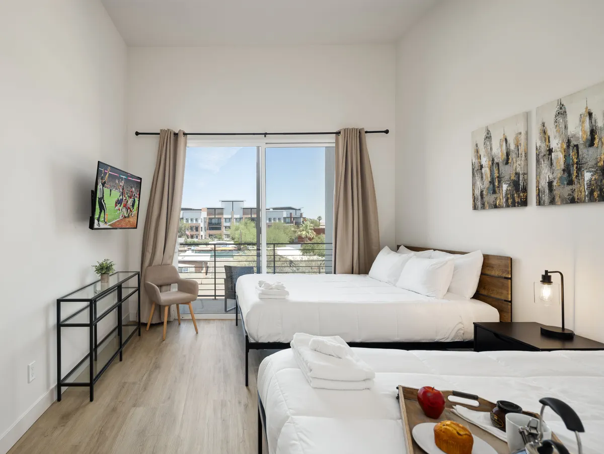 A spacious bedroom with two beds dressed in white bedding, a large window letting in natural light, a television mounted on the wall, and artwork above the beds. A side table with snacks adds to the welcoming, lived-in feel.