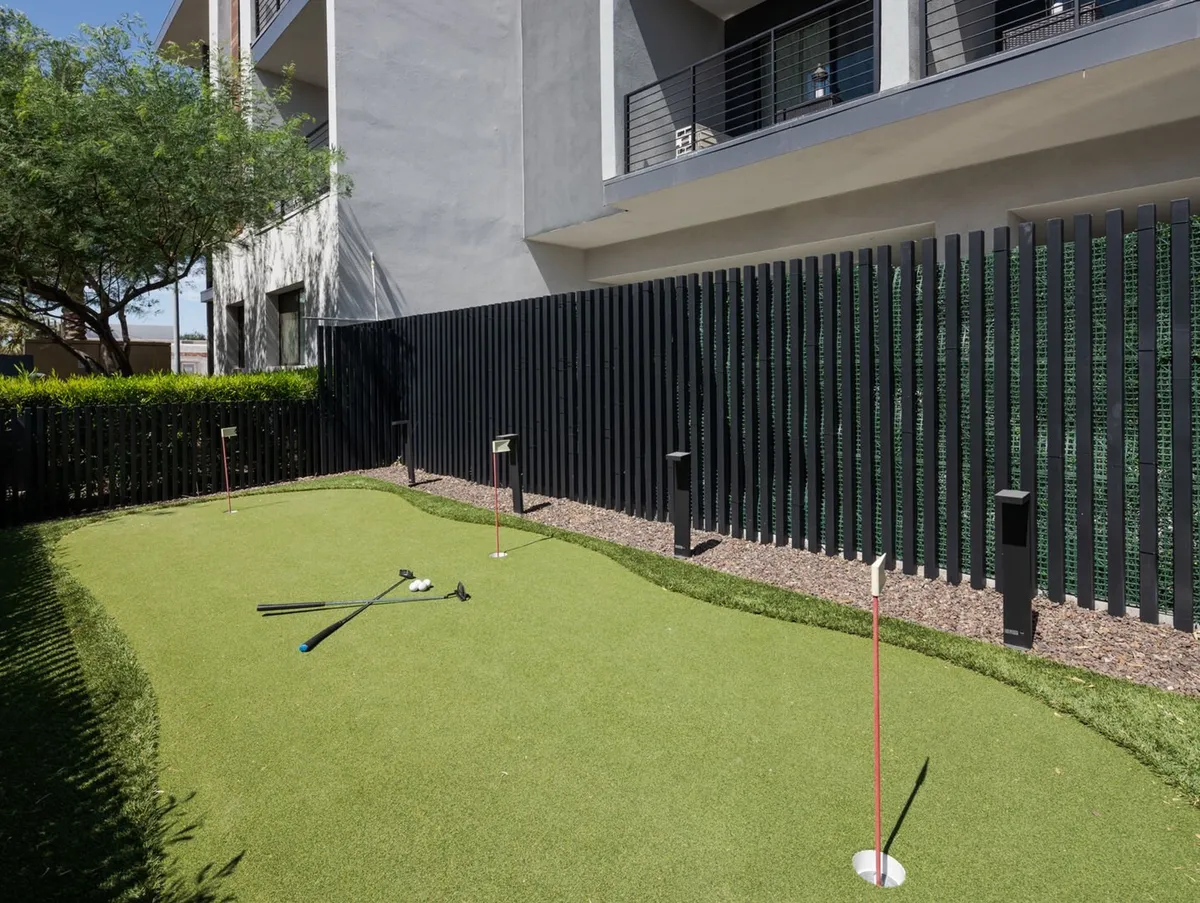 A sunlit outdoor putting green bordered by modern architecture and landscaped greenery, with golf clubs set up for practice and social play. The space exudes a sense of fun and relaxation, designed for friendly competition and group enjoyment.