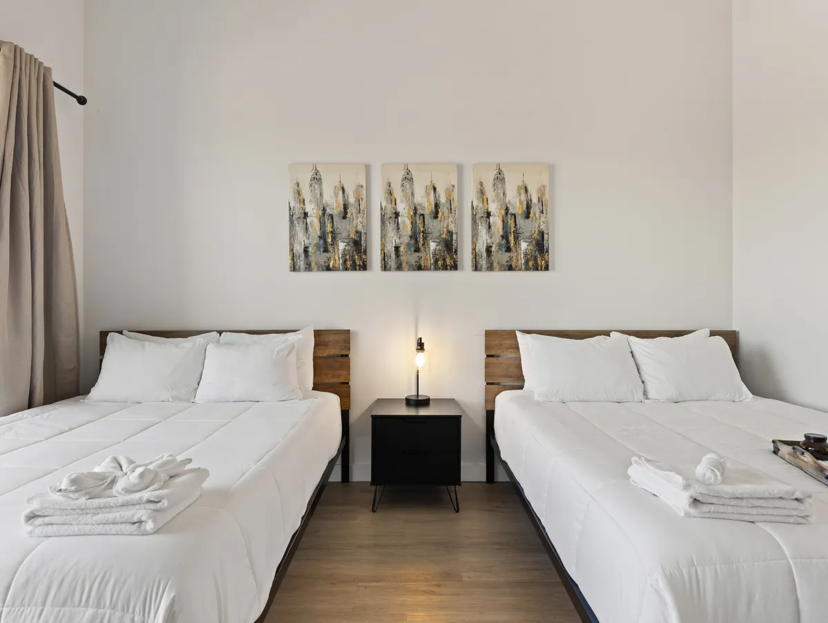 This image features two beds with crisp white linens, accented by abstract artwork on the wall and a nightstand with a lamp between the beds. The overall ambiance is serene and inviting, emphasizing comfort and relaxation—ideal for a restful bedroom retreat.