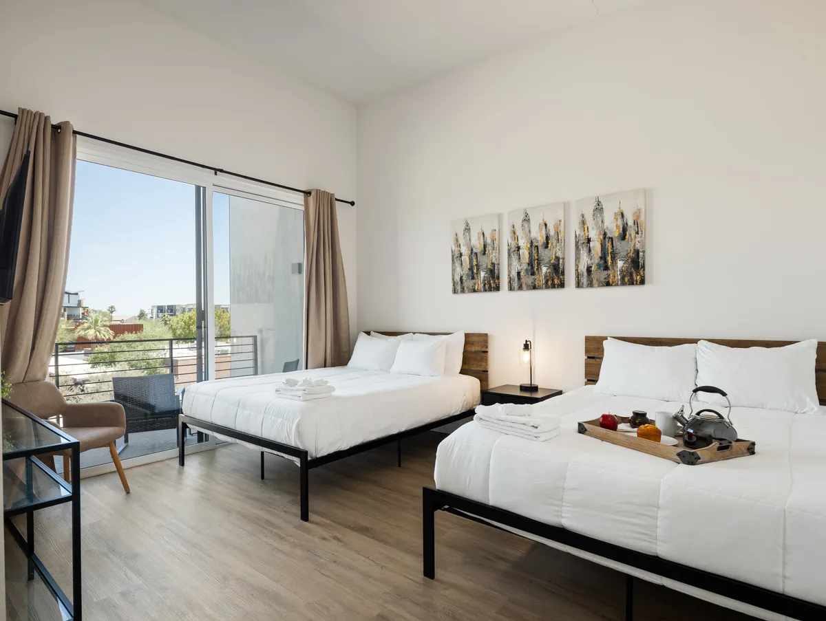 The image showcases a serene bedroom interior featuring two stylishly made beds, contemporary wall art, and a welcoming coffee tray arranged for guests. Natural light pours into the room, casting a warm, inviting glow, while the decor suggests both comfort and thoughtful design.