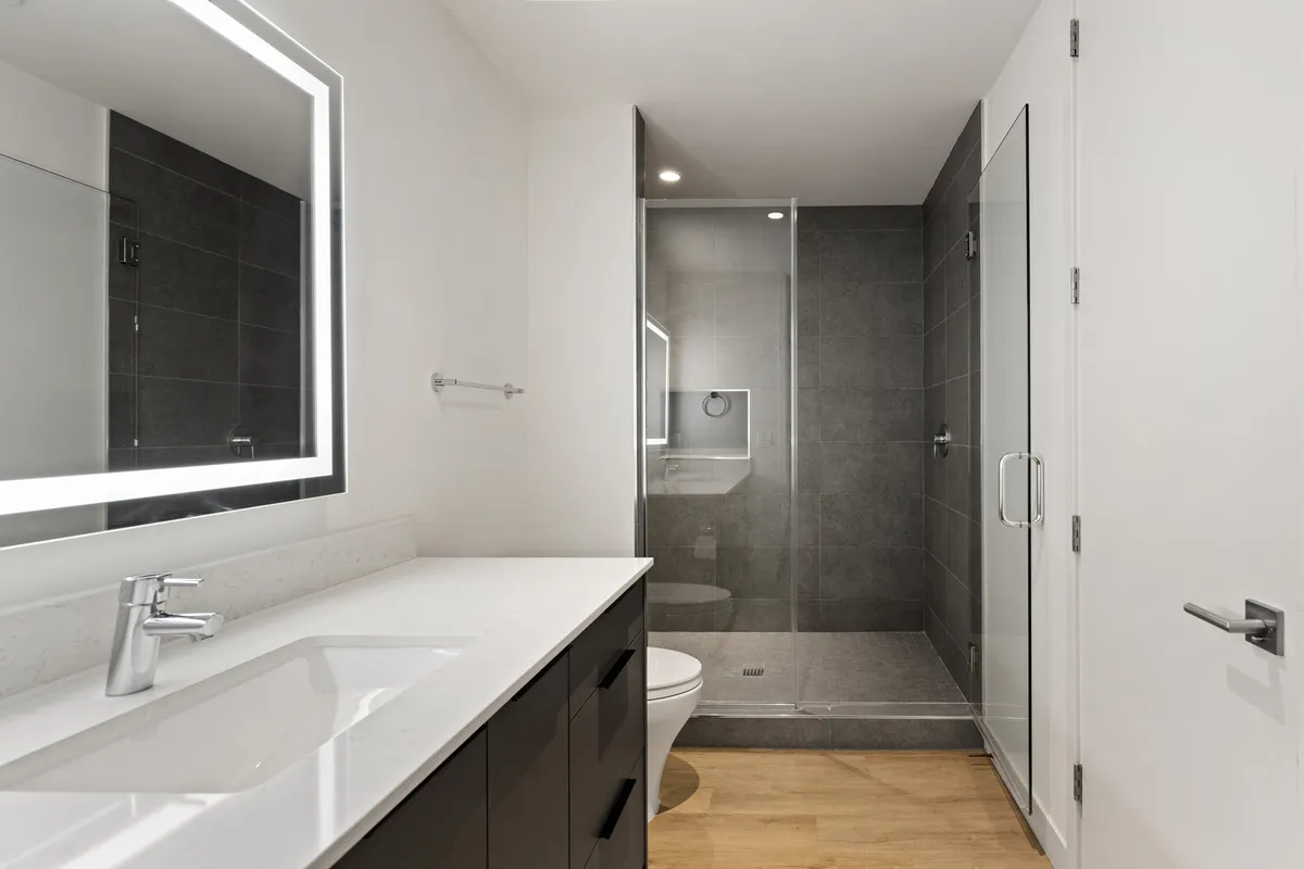 A modern ensuite bathroom featuring a vanity with sink, large mirror, and a glass shower enclosure. The tranquil space is filled with gentle morning light, evoking a spa-like sense of relaxation and privacy.